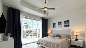 A modern bedroom with a large bed, a ceiling fan, and a view of a pool and palm trees through the glass door. - Villa Elements - new, on water, pool, spa, modern (Cape Coral)
