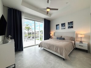 A modern bedroom with a large bed, a ceiling fan, and a view of a pool and palm trees through the glass door. - Villa Elements - new, on water, pool, spa, modern (Cape Coral)