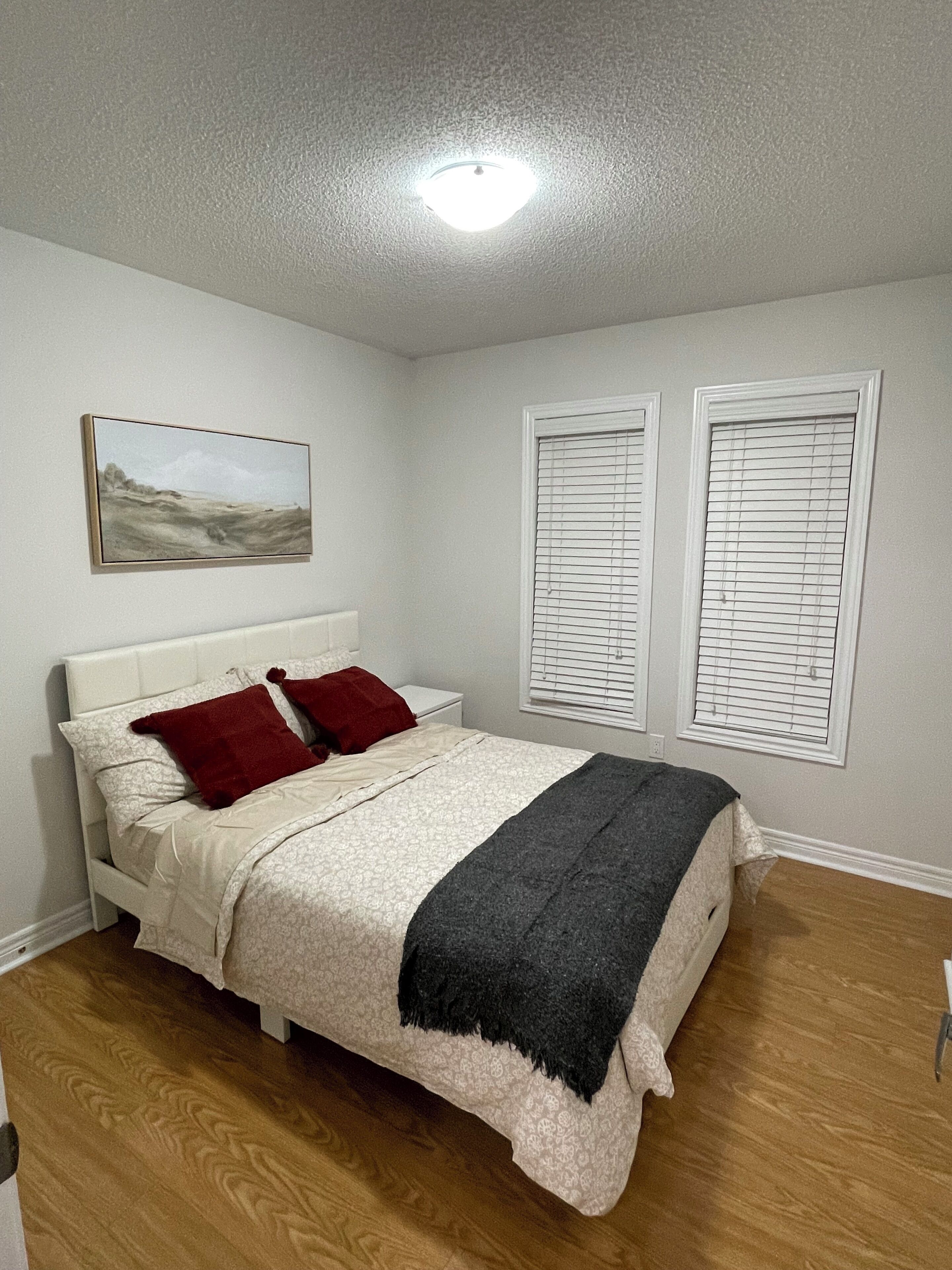 A bedroom with a bed, a painting on the wall, and two windows with blinds.