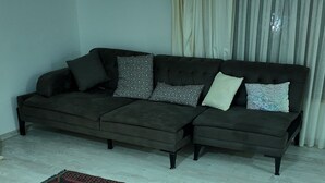 A living room with a dark-colored sofa, patterned cushions, a large patterned rug, and a window with curtains. - KARYA OTEL (Çandır)