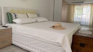 A neatly made bed with a white bedspread and a hat, a bedside table with a remote control, a wooden nightstand, and a wardrobe. - KARYA OTEL (Çandır)