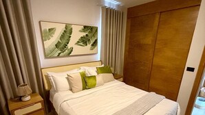 A neatly made bed with white linens and a green throw, a wooden nightstand with a lamp, a large painting of leaves on the wall, and a wooden wardrobe. - Poniente 201 – Tropical Garden Apartment with Pools near Dominicus Beach (Dominicus)