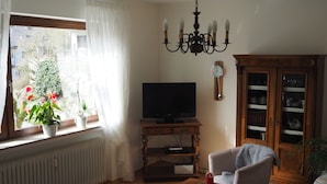 A living room with a television, a chandelier, a cabinet with glass doors, and a window with curtains. - Vacation apartment Haus Junk am Lieserpfad (Daun)