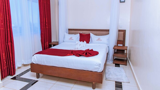 A hotel room with a wooden bed, white bedding, red accents, and a view of the outdoors. - Hotel OCCAZIA Homa Bay (Homa Bay)