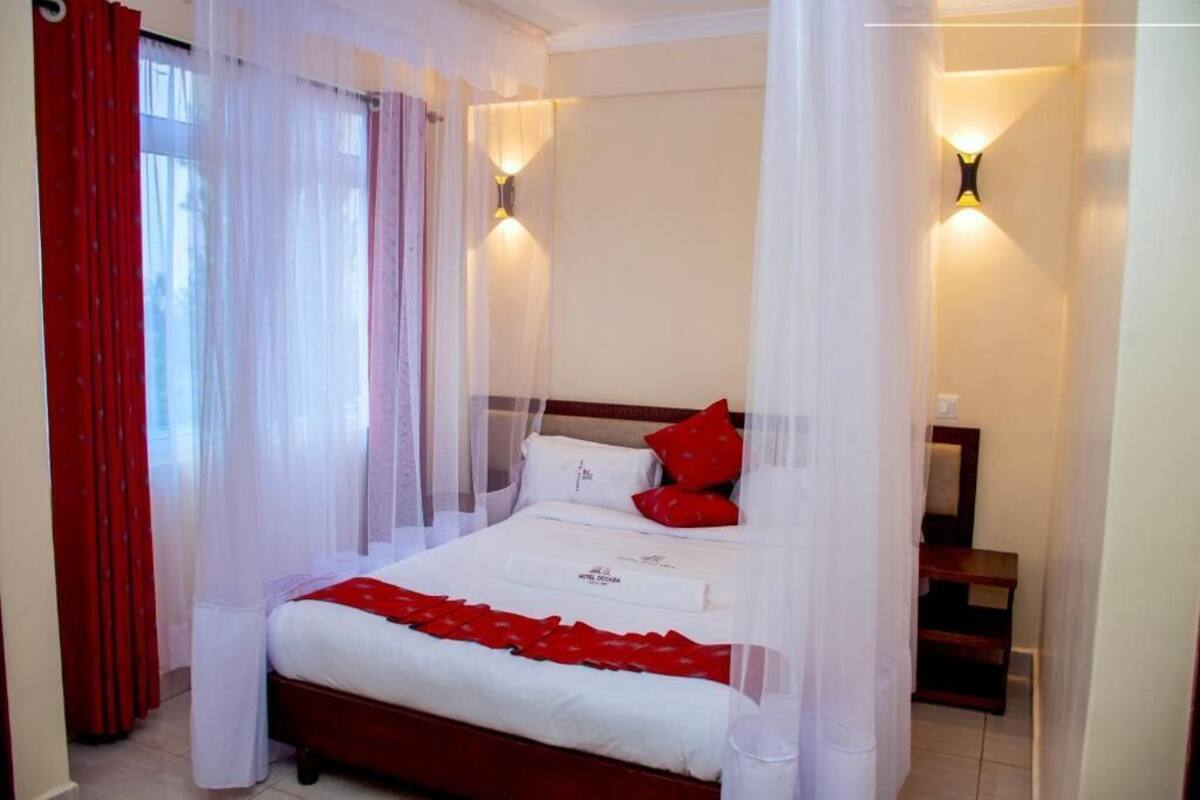 A hotel room with a four-poster bed, white bedding, red pillows, and a canopy. There are two wall-mounted lights on either side of the bed.