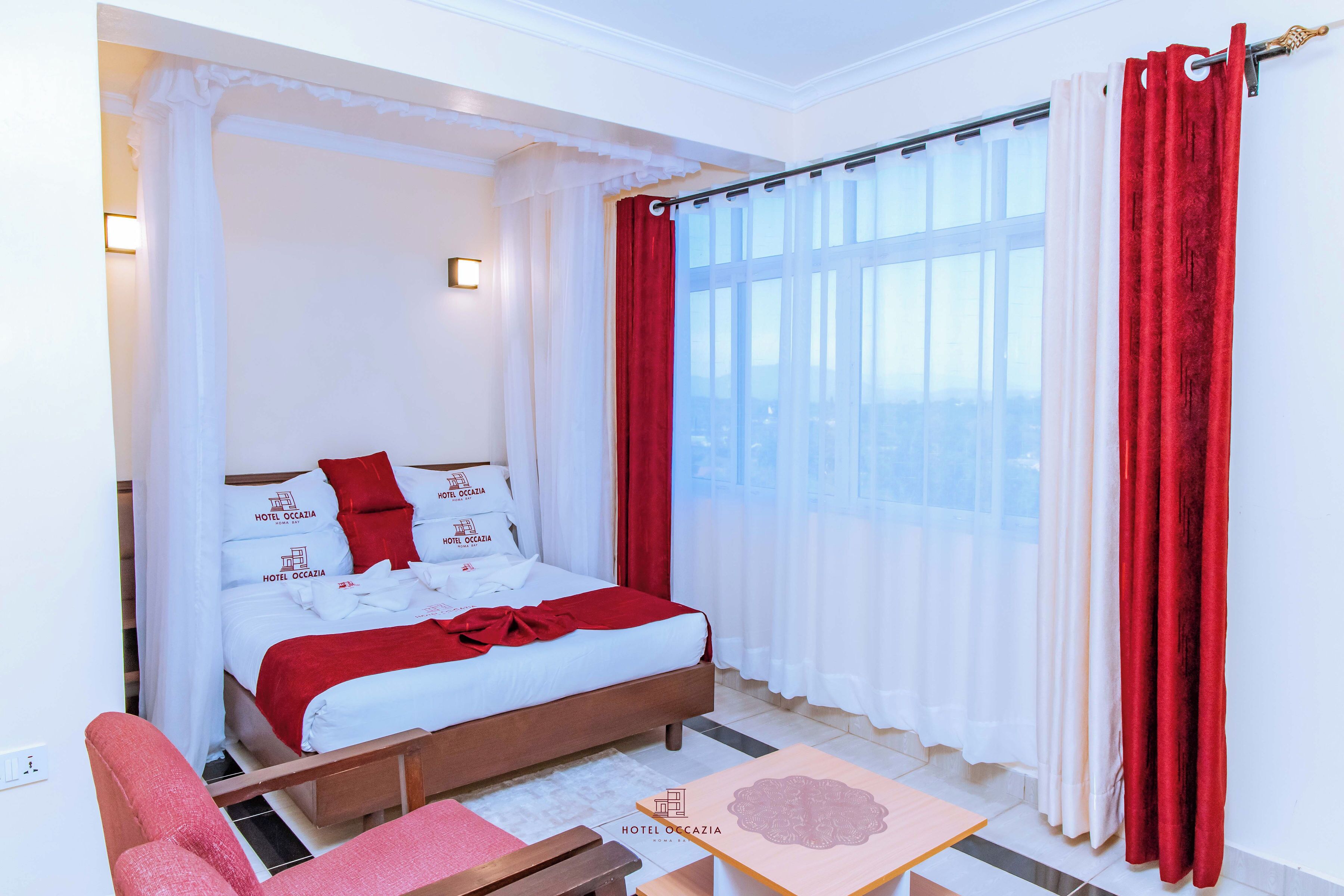 Quarto de hotel com cama, mesa de cabeceira, cadeira e janela com cortinas.