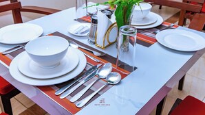 A dining table set with white plates, silverware, and a glass of water. There is a vase with flowers and a salt and pepper shaker on the table. - Hotel OCCAZIA Homa Bay (Homa Bay)