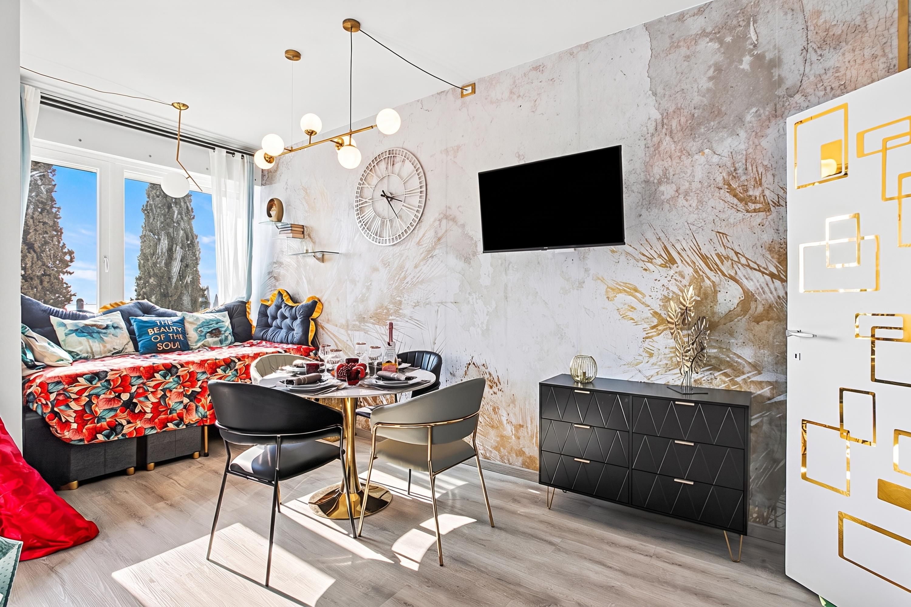 A modern living room with a dining area, a large window, a flat-screen TV mounted on the wall, and a decorative wall piece.