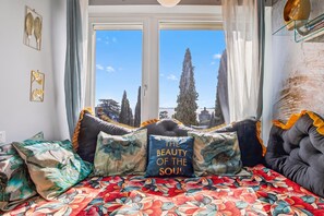 Une chambre avec un lit, une fenêtre donnant sur des arbres et un immeuble, et un coussin décoratif orné d’un motif textuel.