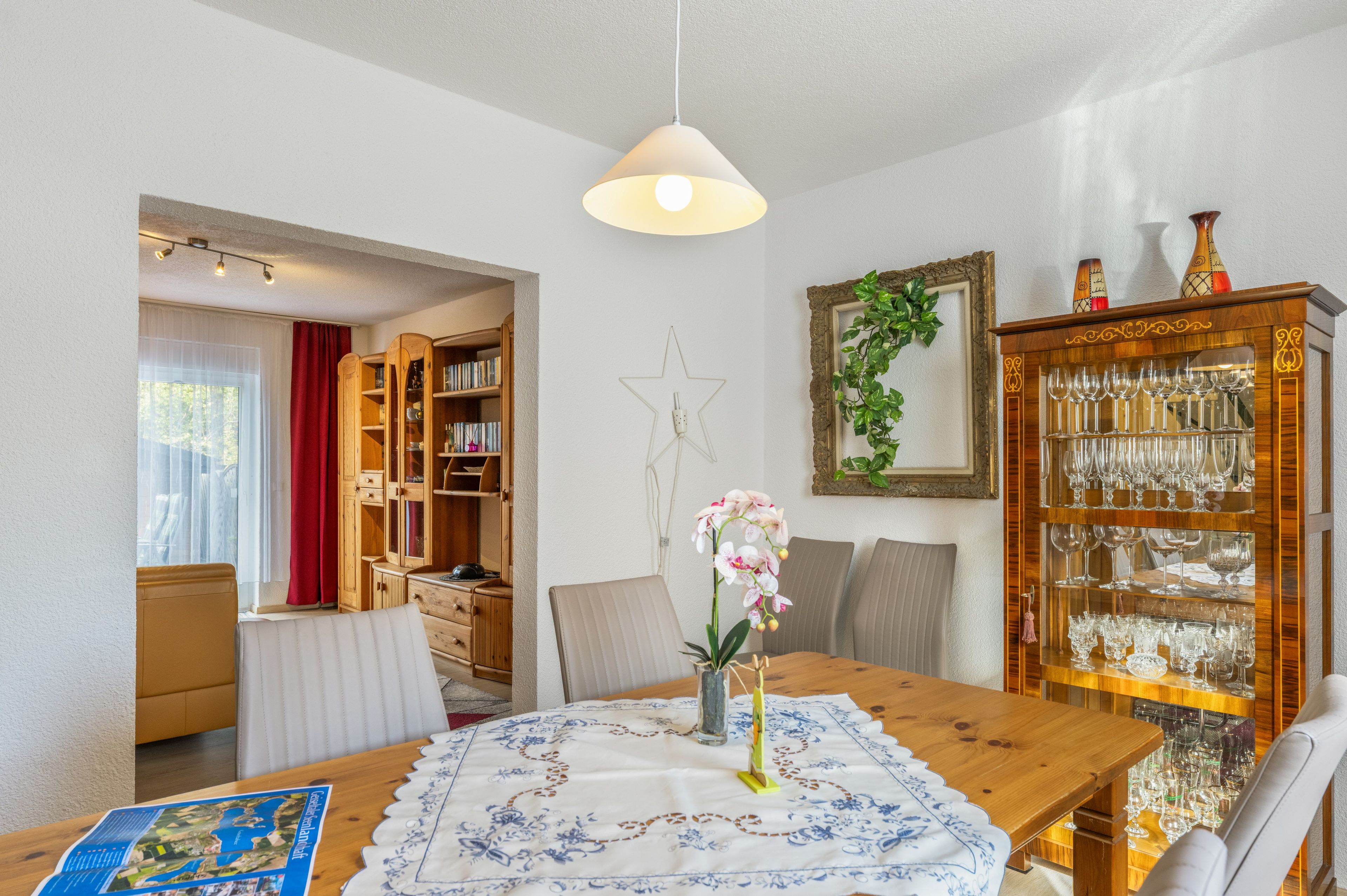 Un comedor con una mesa de madera, sillas, un jarrón con flores, un mapa y una estantería.