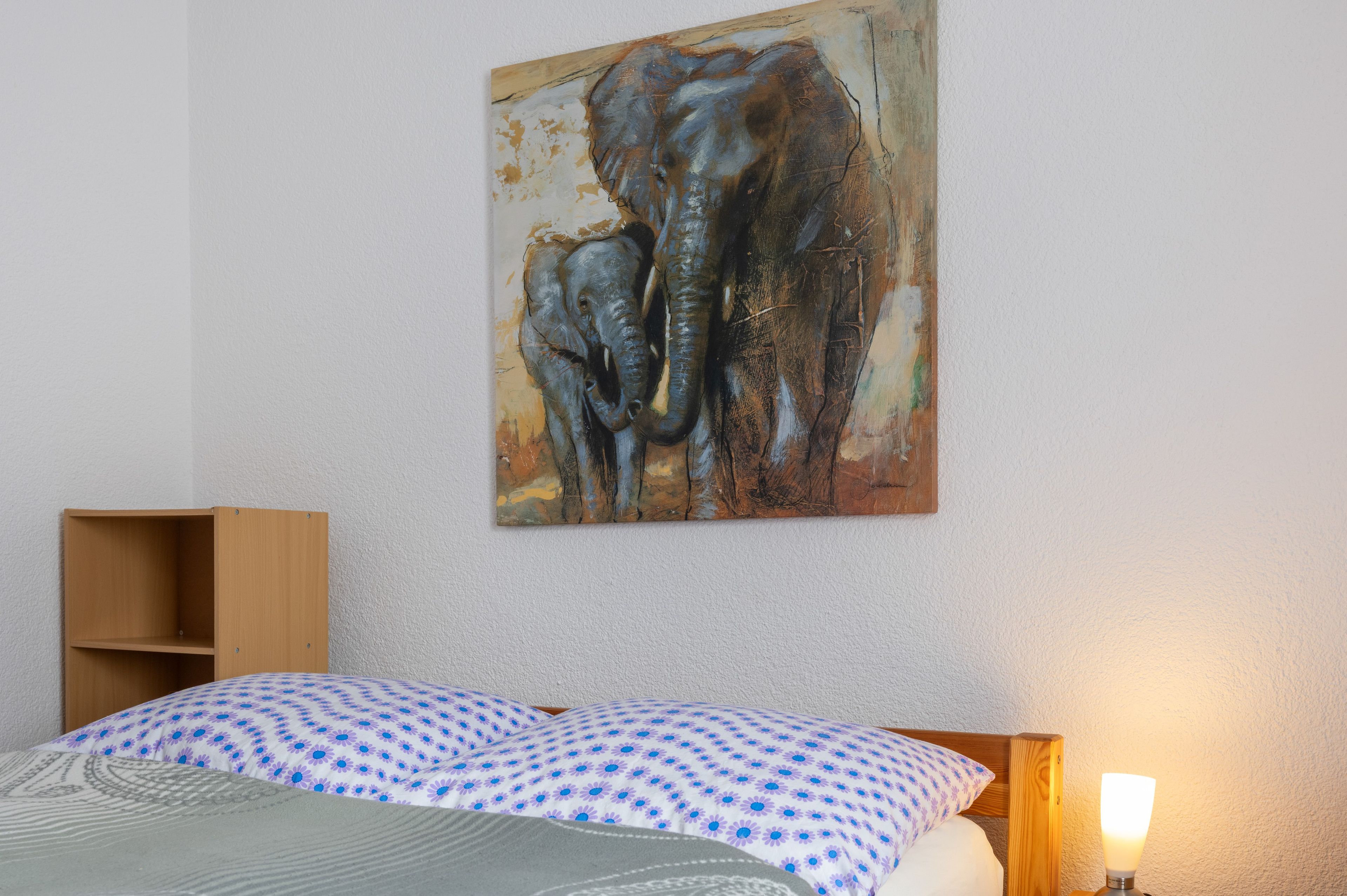 Un dormitorio con cama, cabecera de madera, mesita de noche y un cuadro colgado en la pared con elefantes.