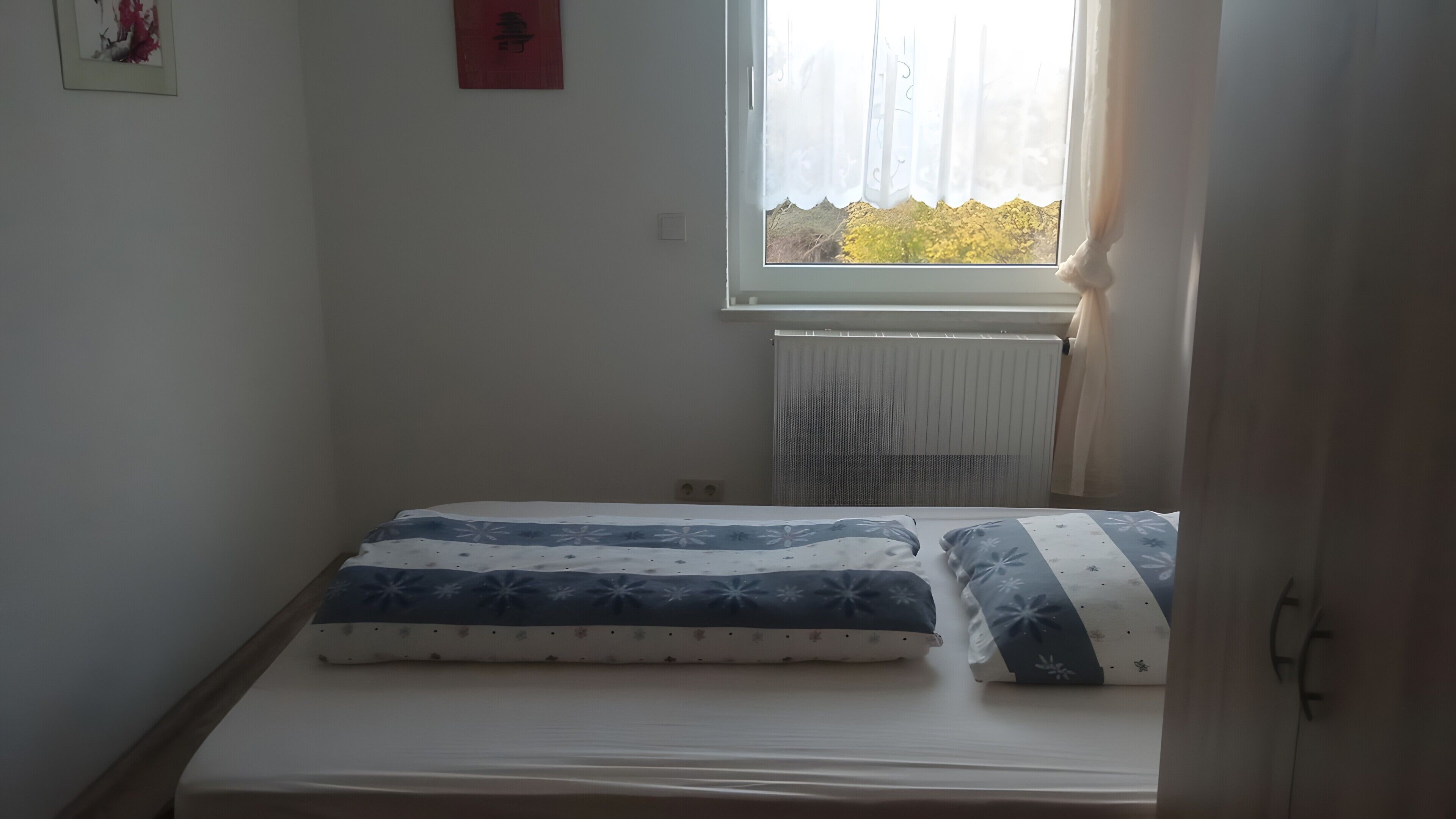 Quarto pequeno com cama de solteiro, janela, radiador e interruptor de luz fixado na parede.