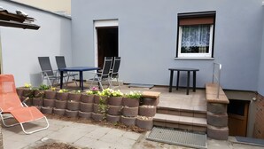 A patio with a table and chairs, a small garden bed with plants, a window with a curtain, and a door.