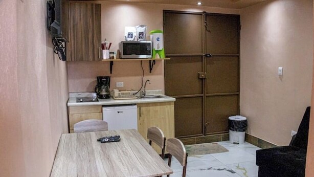 Cocina compacta con microondas, cafetera y un refrigerador pequeño. Hay una mesa de comedor con sillas y un sofá negro.