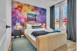 Un dormitorio con una pared temática de galaxia, una cama con ropa de cama blanca, dos mesitas de noche con lámparas y vistas a una gasolinera por la ventana.