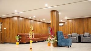 A lobby with a brown leather sofa, a blue armchair, and a grey sofa. There are potted plants and a decorative pillar. - Hotel Orcella (Tirupati)