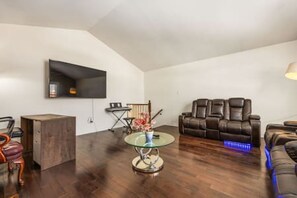 Un salón amplio con una televisión de pantalla plana montada en la pared, una mesa de centro de vidrio, un sofá de cuero y un pequeño armario de madera.