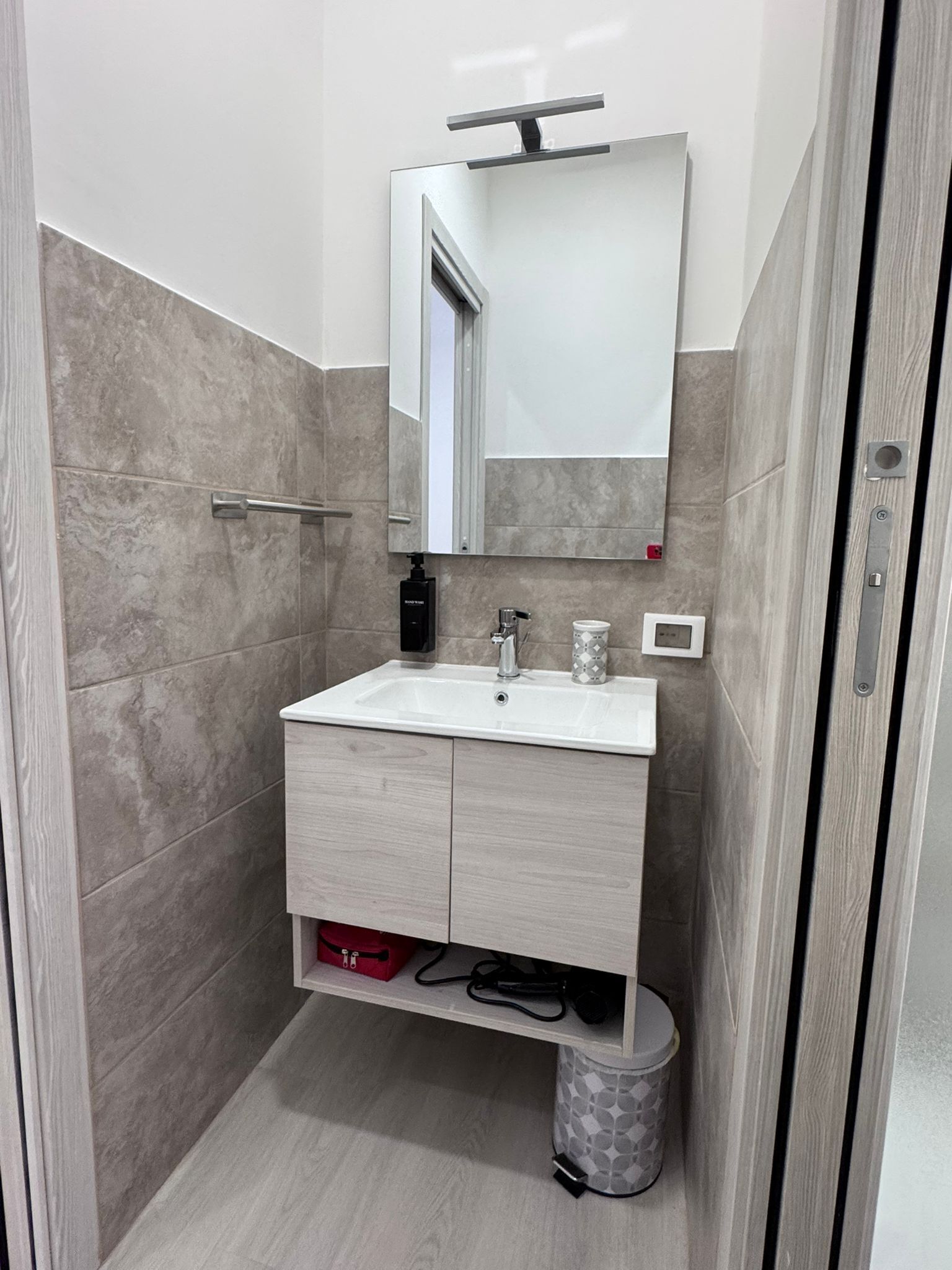 Une salle de bain avec un lavabo blanc, un miroir, un distributeur de serviettes et une poubelle.