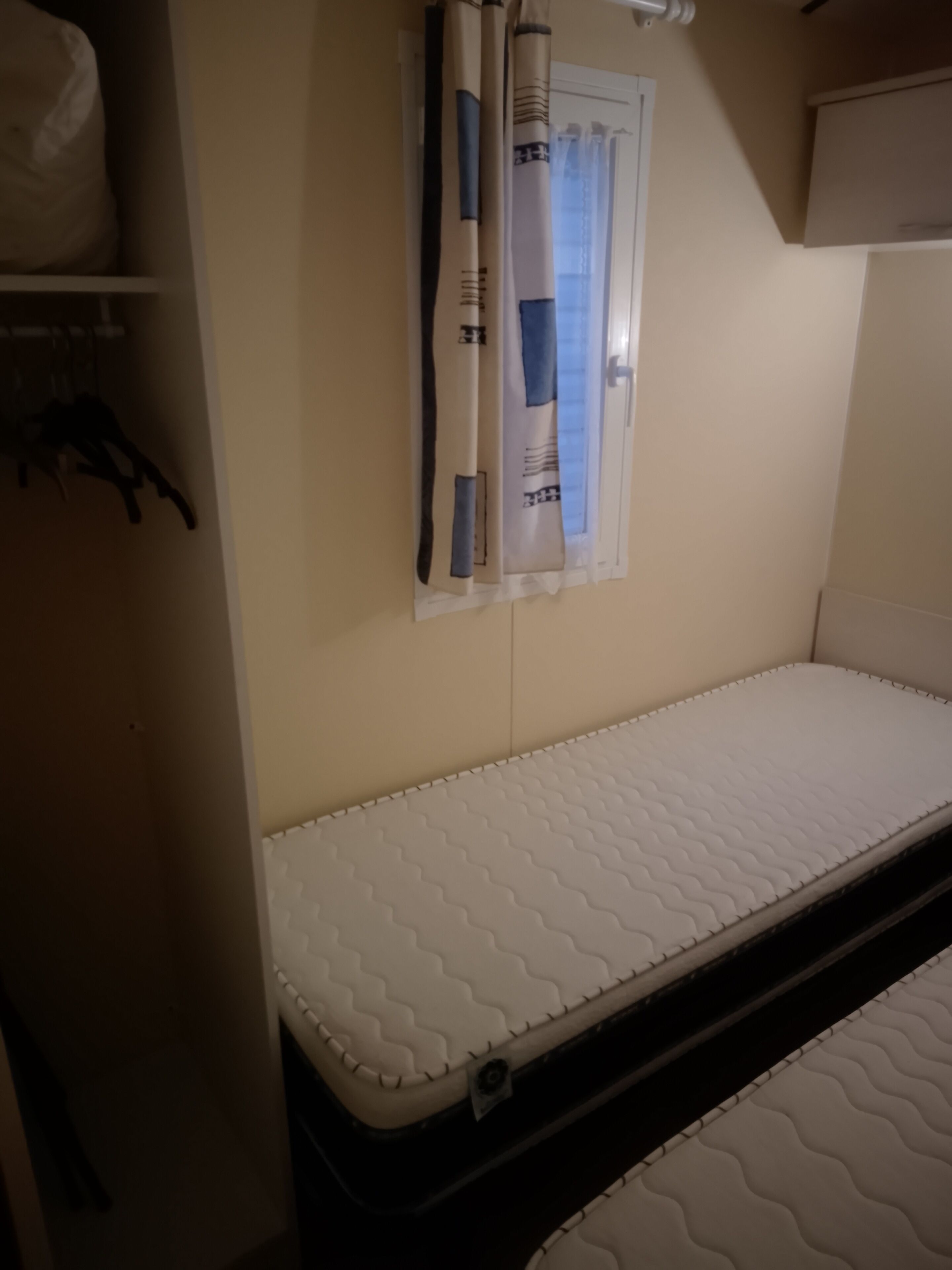 A small, single-bed room with a white mattress, a window with curtains, and a shelf with hanging items.