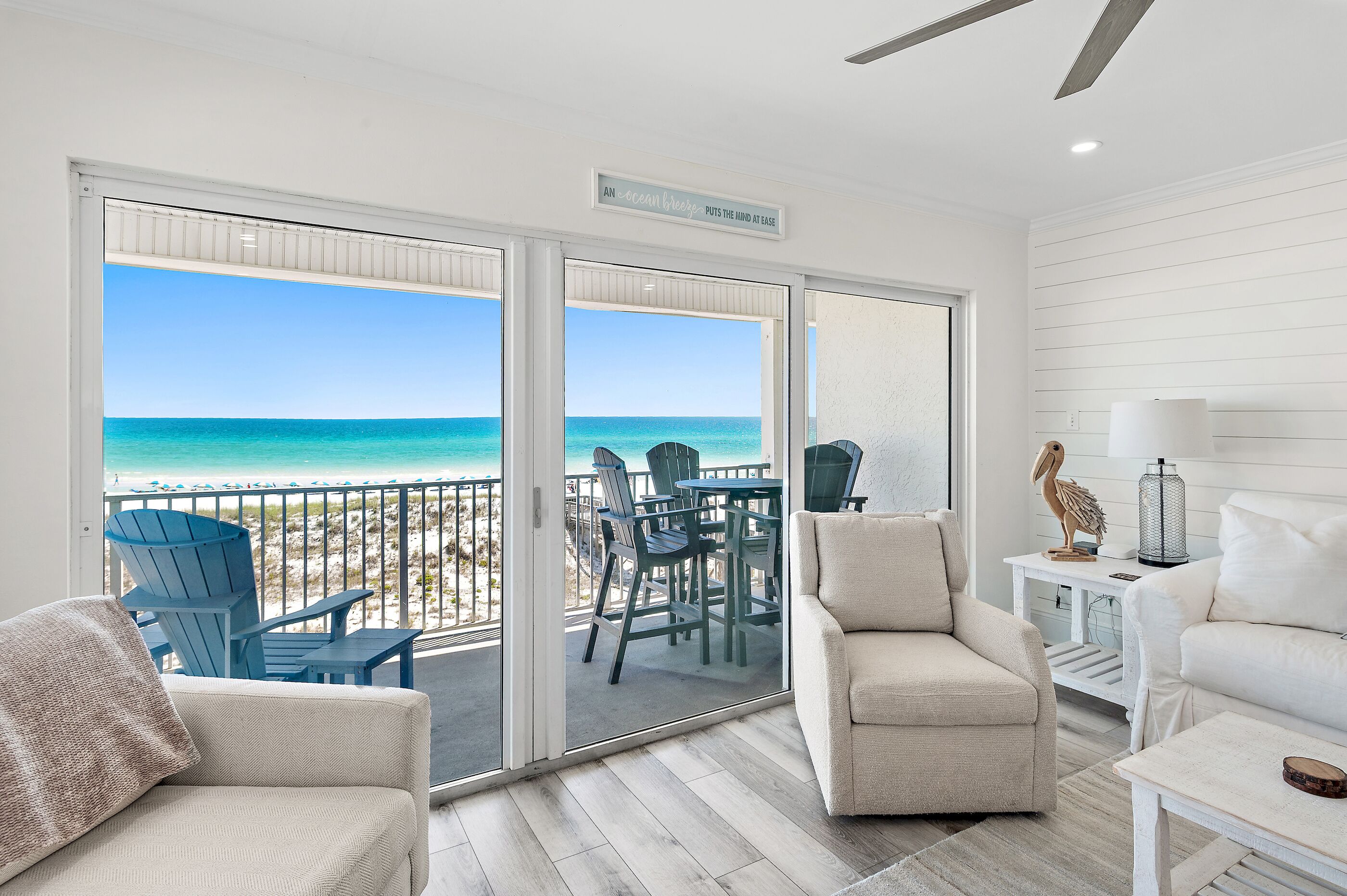 Una habitación con vistas a la playa, una puerta corrediza de vidrio, un sofá blanco, una silla azul y una mesita con una lámpara.