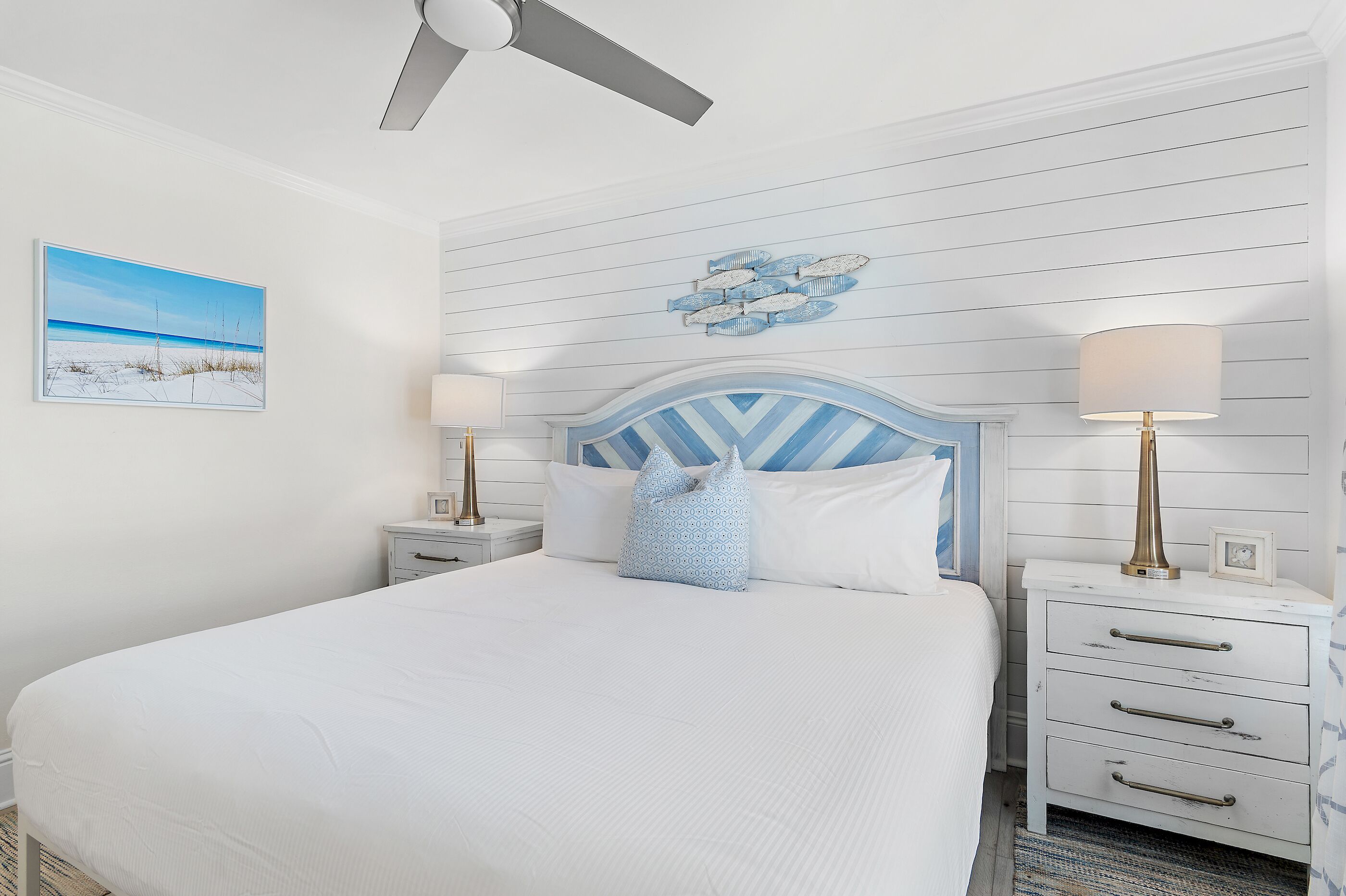 A bedroom with a bed, two bedside tables, a ceiling fan, a wall-mounted artwork, and a decorative wall panel.