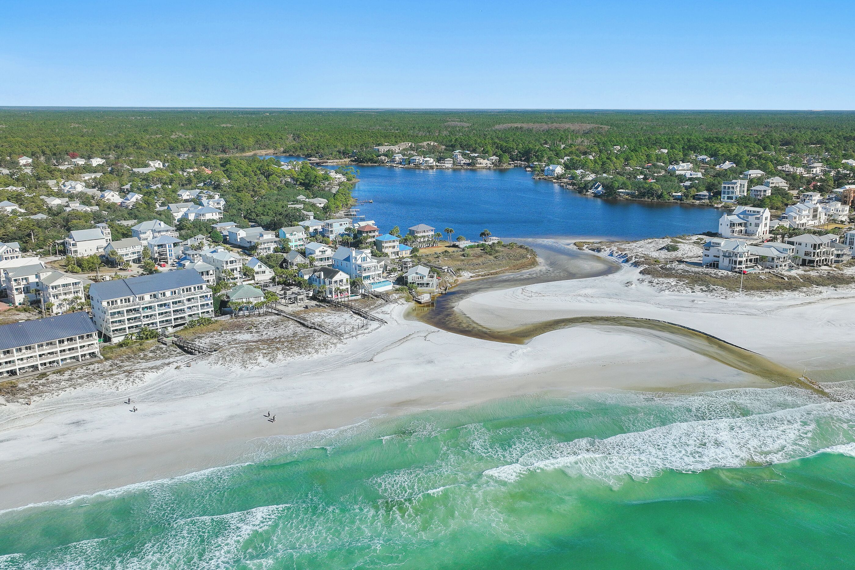 An aerial view of a coastal residential area with beachfront properties, a curving road, and a clear blue sky.