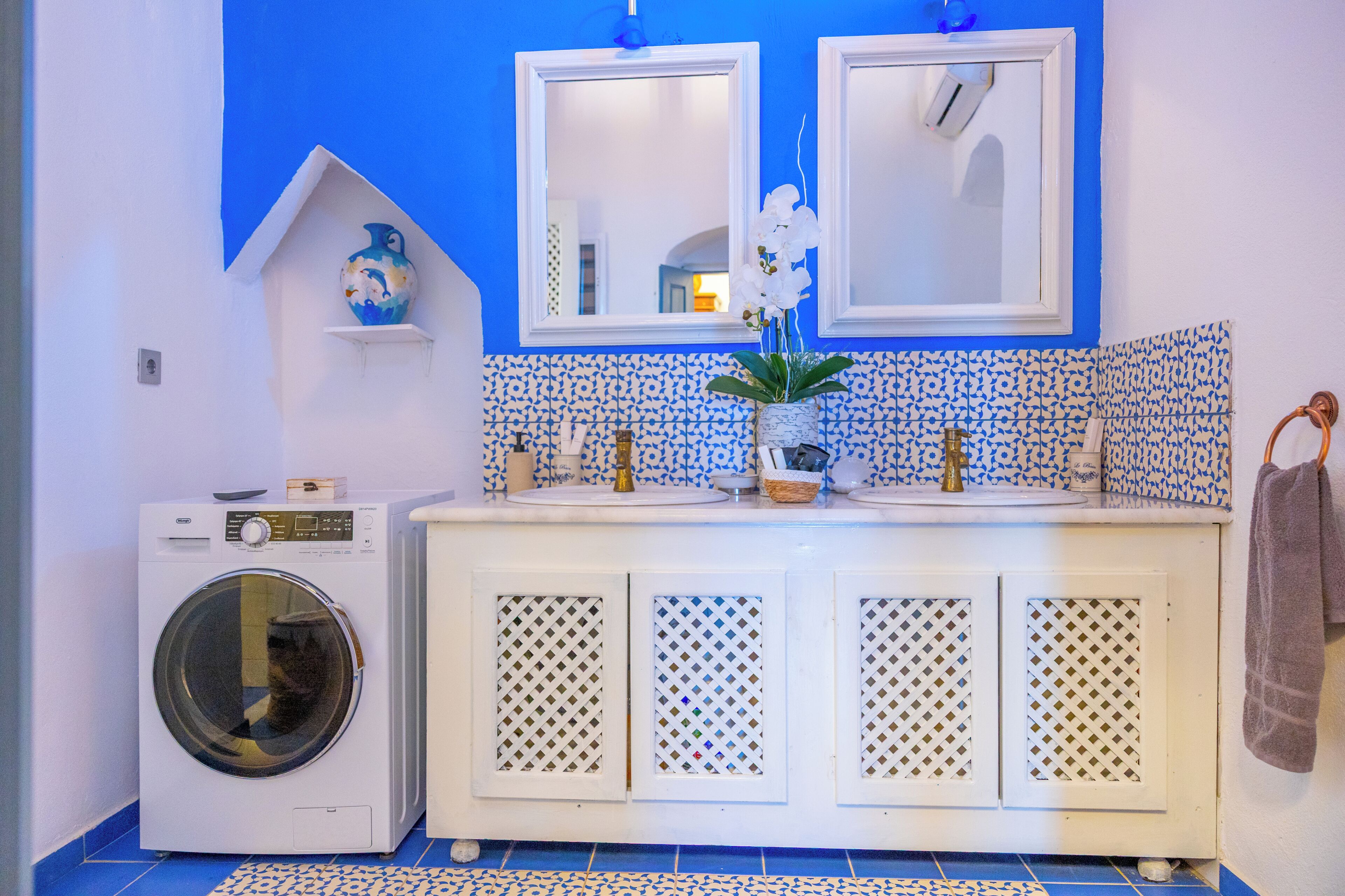 Un baño con lavadora, dos espejos, un lavamanos con un florero y baldosas estampadas.