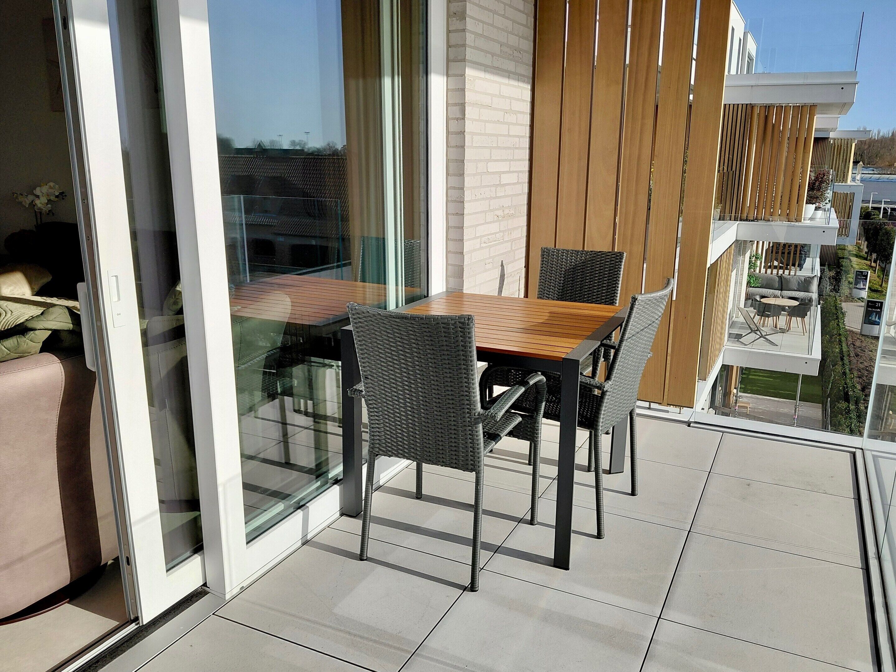 Ein Balkon mit einem Holztisch, Korbstühlen, einer Glastür und Blick auf ein Gebäude mit weißer Backsteinfassade.