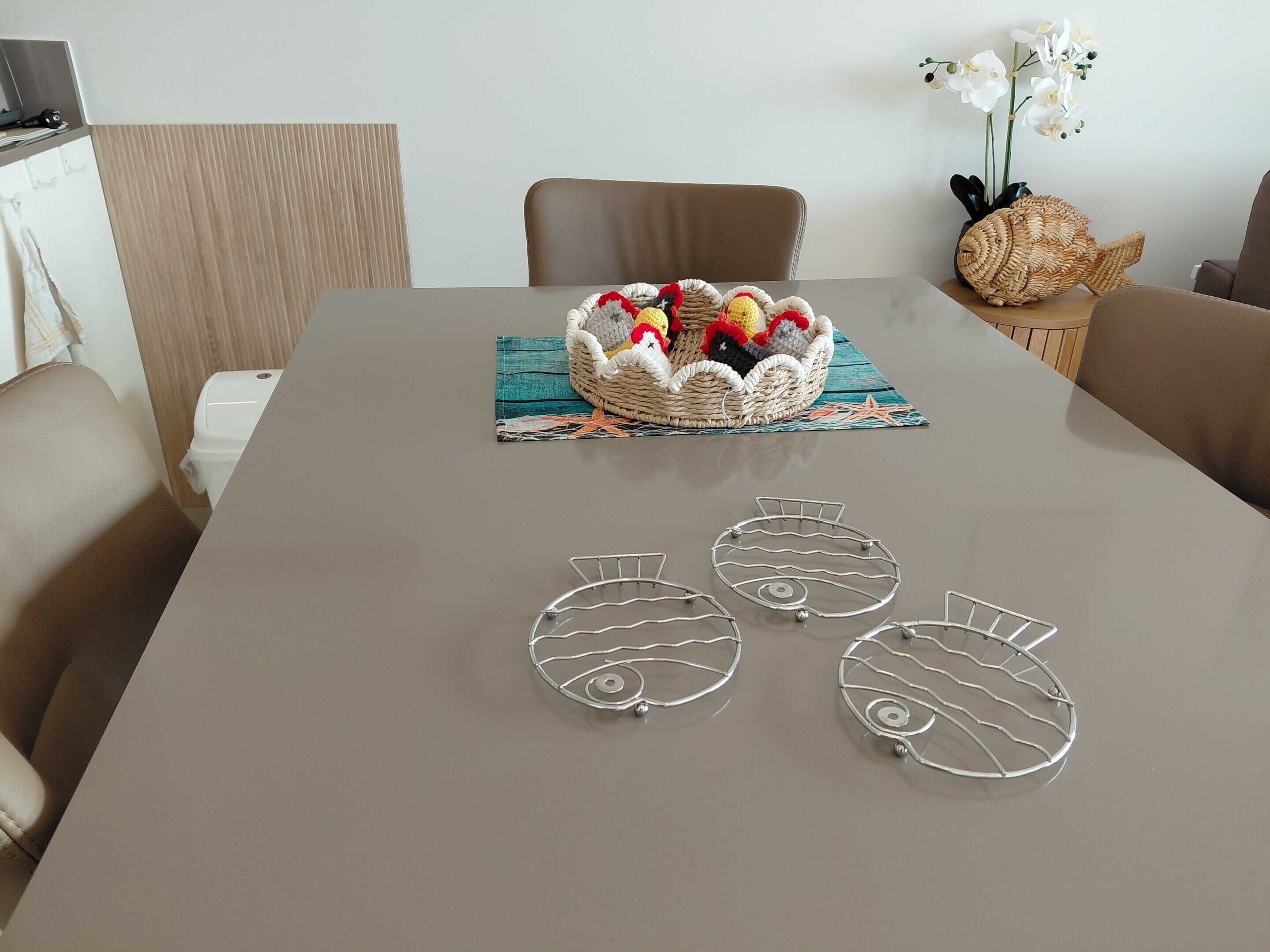 A dining table with a woven basket centerpiece, three wire fish-shaped coasters, and a vase with white flowers.
