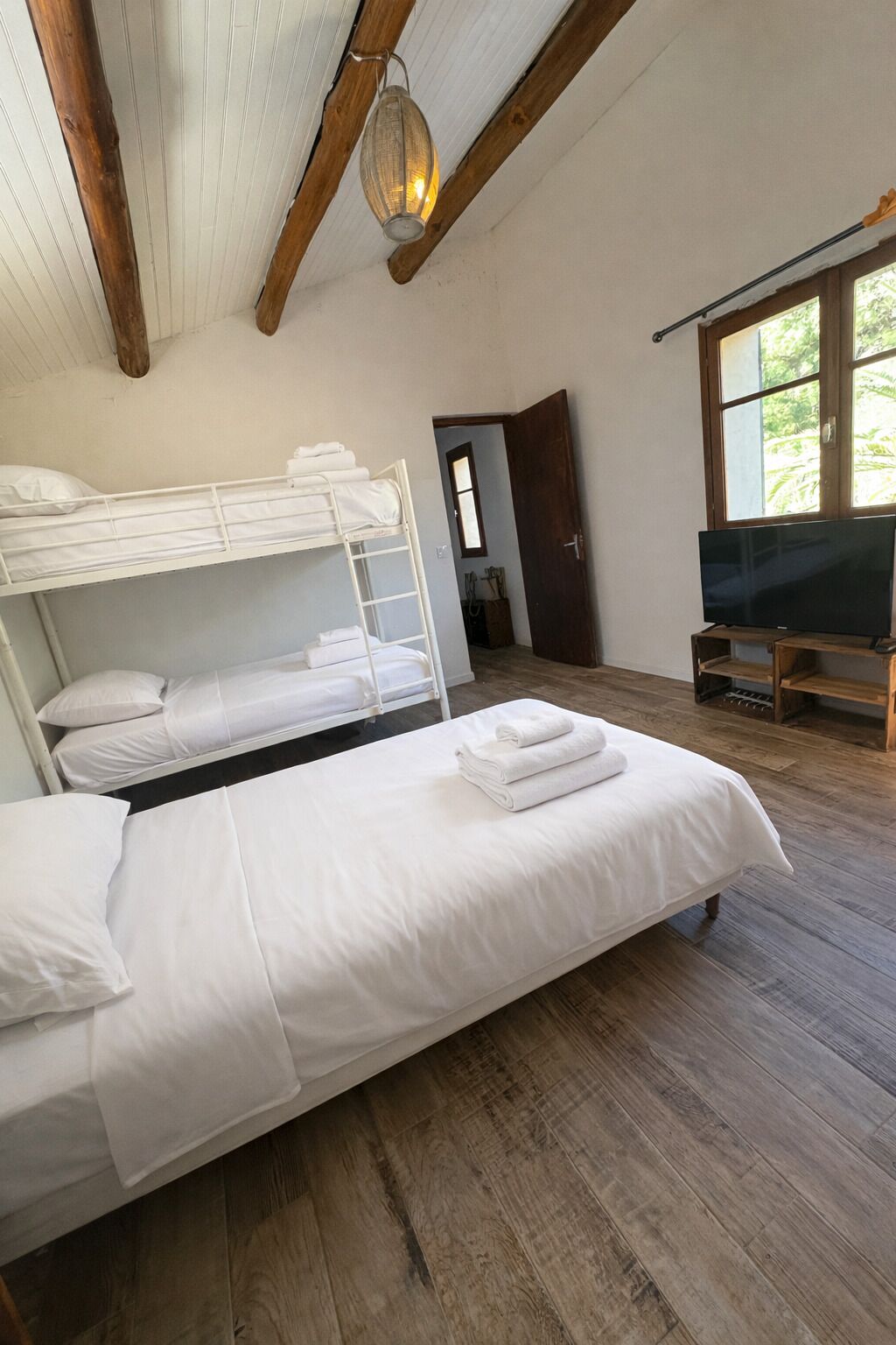 Ein Zimmer mit einem Etagenbett, einem Doppelbett, Holzboden, einem Fernseher und einem Fenster mit Blick auf Grünflächen.