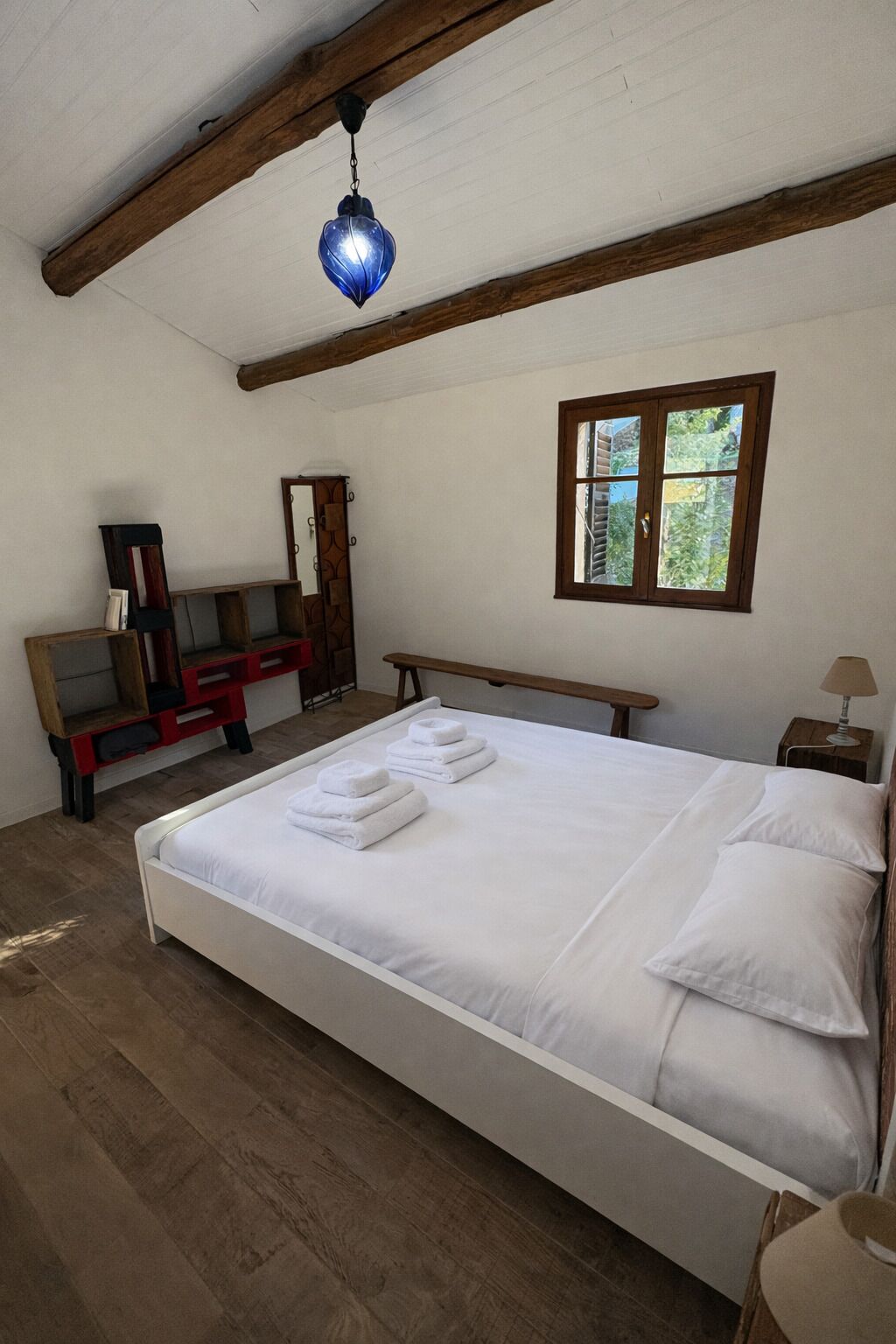 A bedroom with a large bed, wooden beams, a window, a desk, and a hanging light fixture.