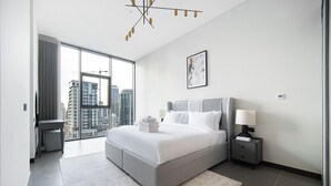 Una camera da letto moderna con un letto grande, un comodino, una lampada da notte e una finestra con vista su alti edifici, illuminata da un lampadario.