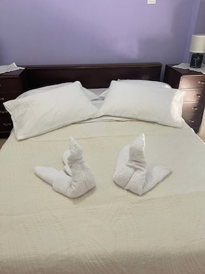 A bed with white linens, two towels folded in a decorative manner, a wooden headboard, and a nightstand with a lamp and a small decorative item.