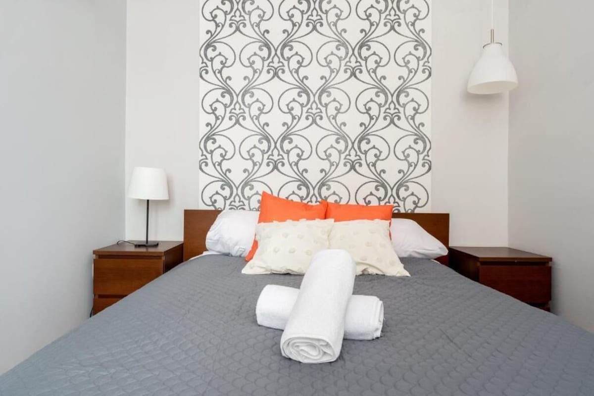 A neatly made bed with a grey quilt, orange and white pillows, and rolled white towels on a wooden nightstand.