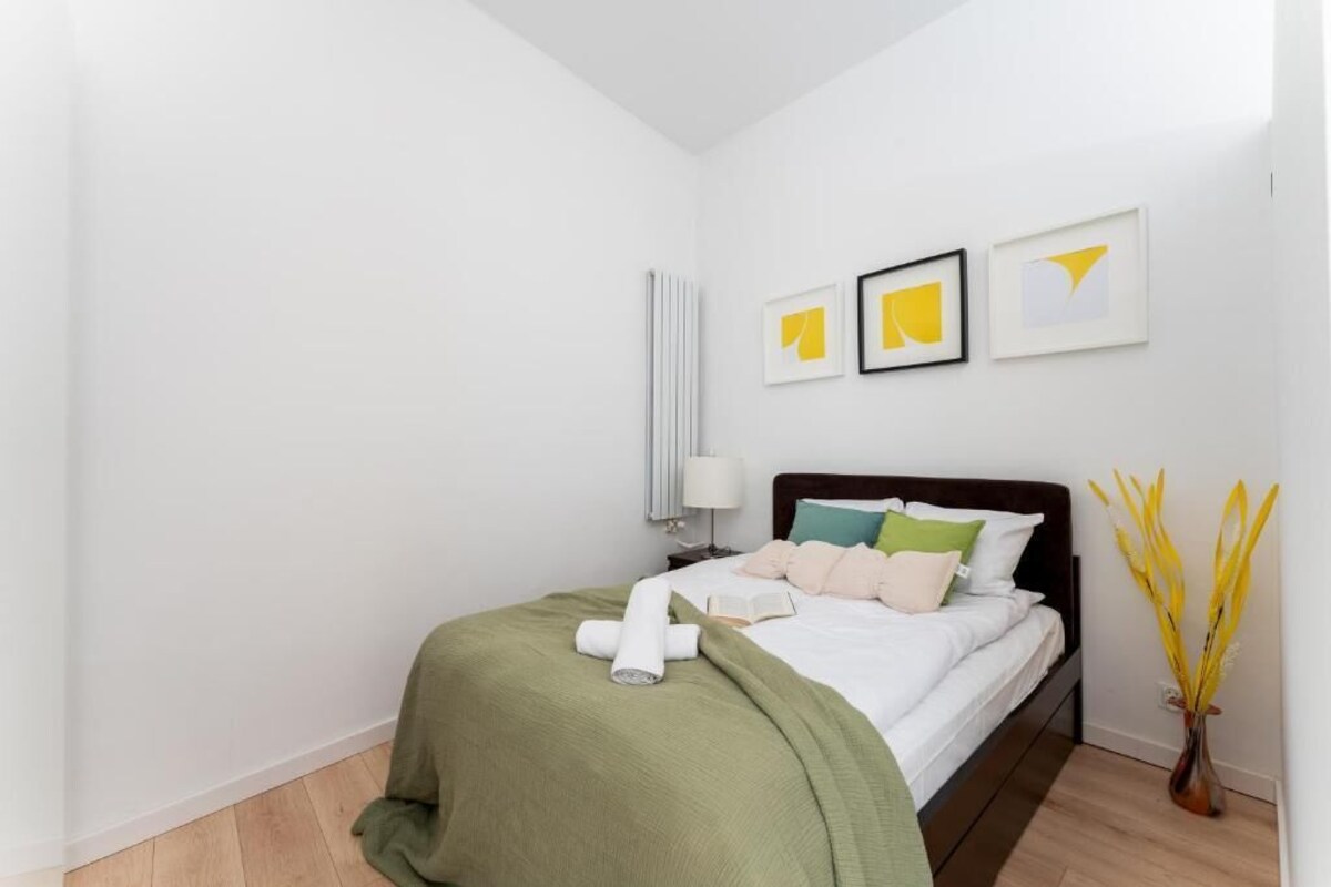 A neatly made bed with a green blanket, a white pillow, and a rolled towel. There are three framed pictures on the wall, a bedside lamp, and a vase with yellow flowers.