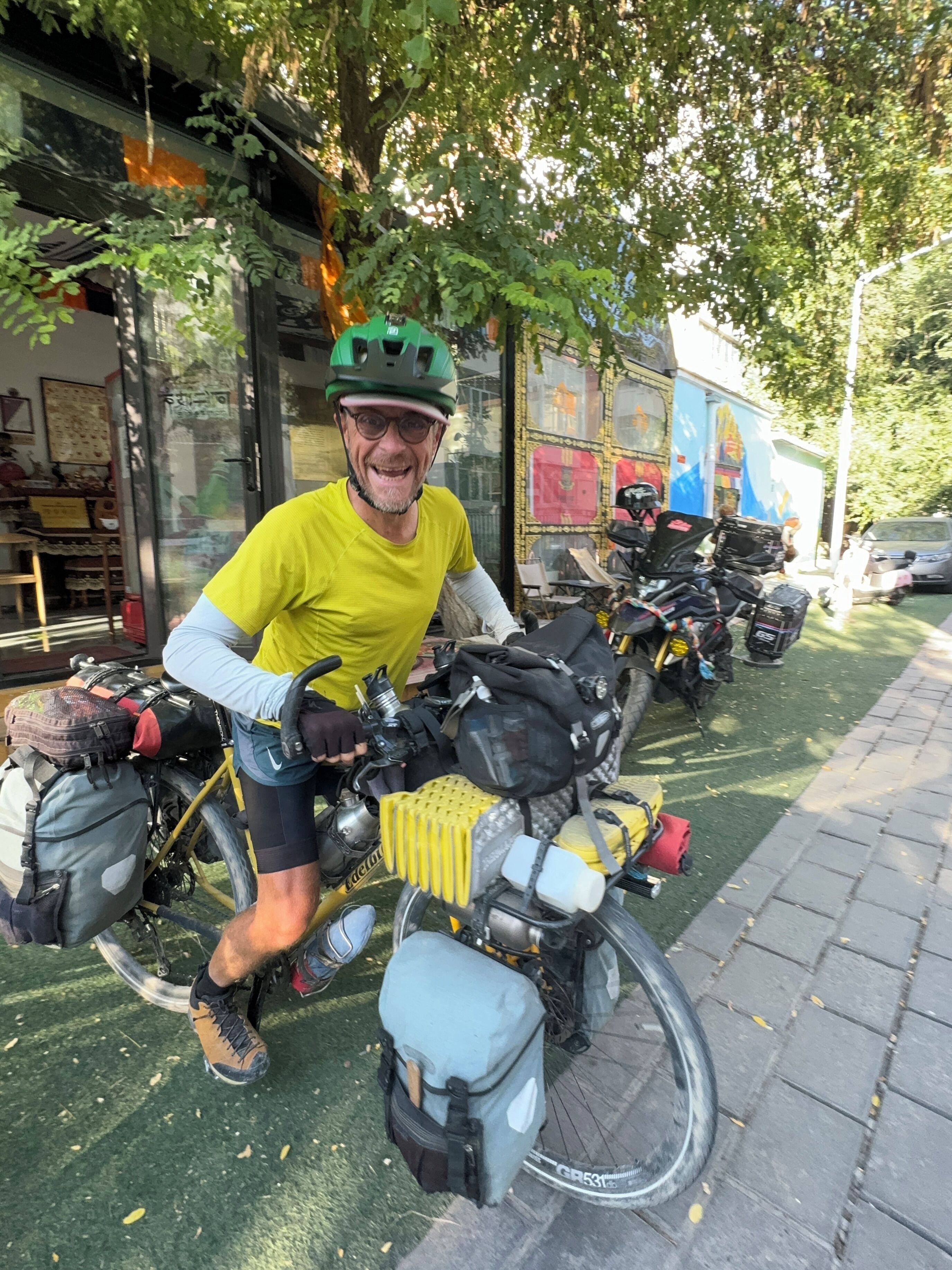 Een persoon op een fiets met meerdere tassen en een helm, geparkeerd op een verhard gebied met bomen en een gebouw op de achtergrond.