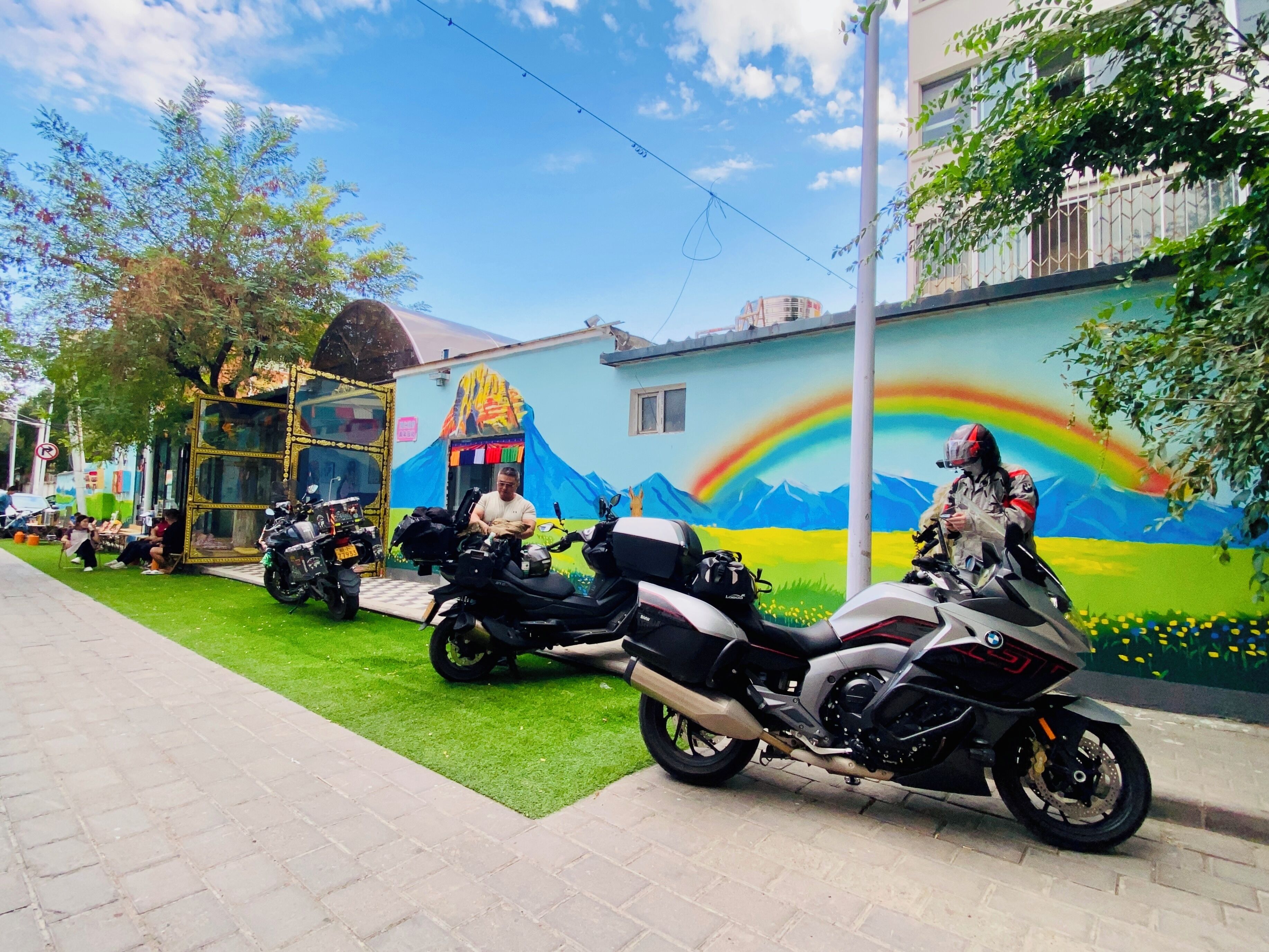 Un trottoir dallé bordé d’une pelouse verdoyante, une fresque représentant un arc-en-ciel et des montagnes, et des motos garées.