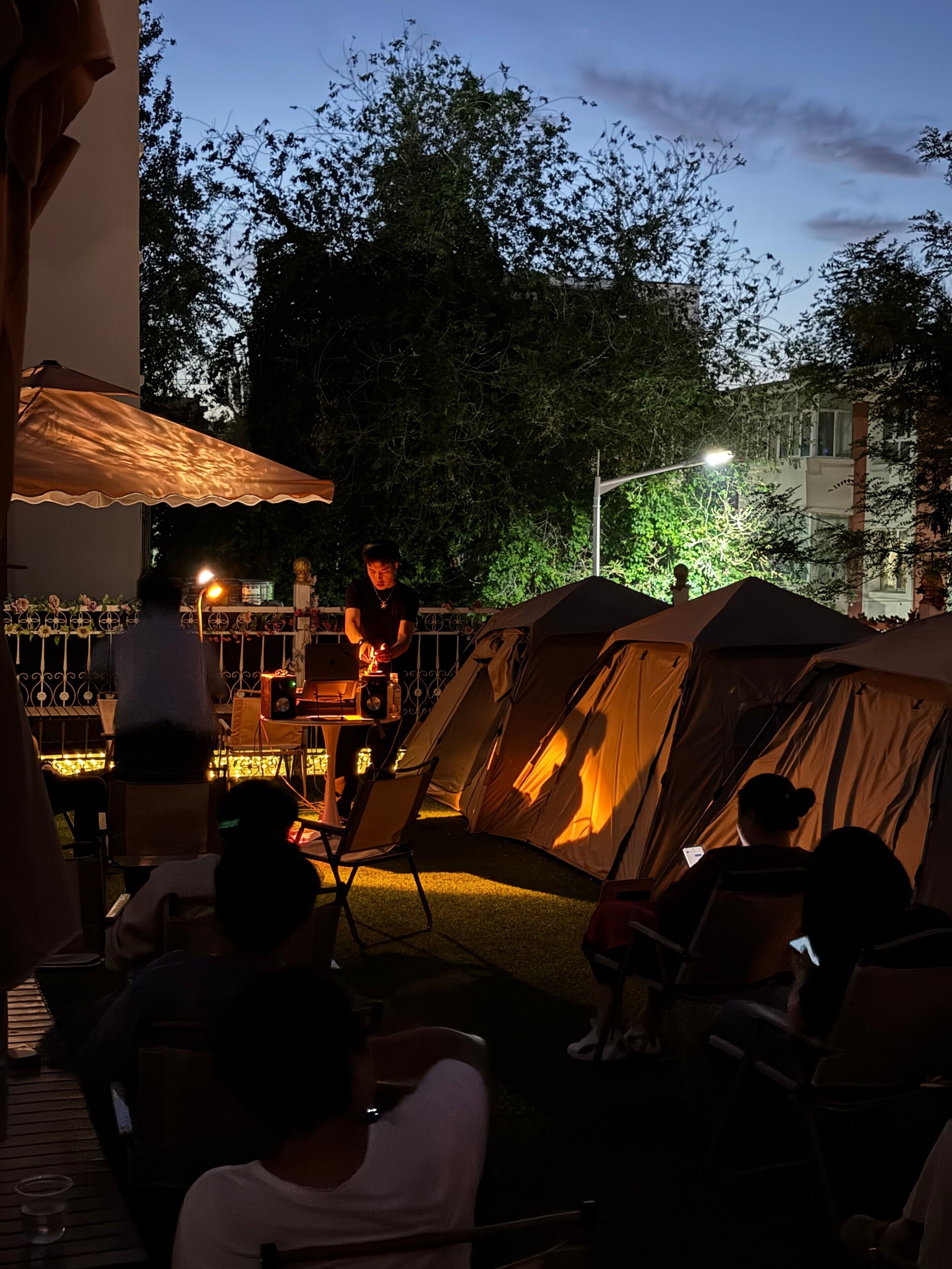 An outdoor dining area with tables and chairs, a tent, and a person cooking on a portable stove.