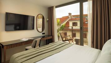 Habitación de hotel con cama, escritorio, silla, televisor y vista a edificios a través del ventanal.