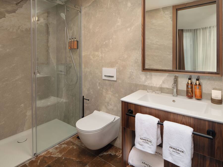 Een badkamer met een douche in een glazen cabine, een wit toilet, een houten wastafelmeubel met een spiegel, en twee handdoeken aan een handdoekhouder.