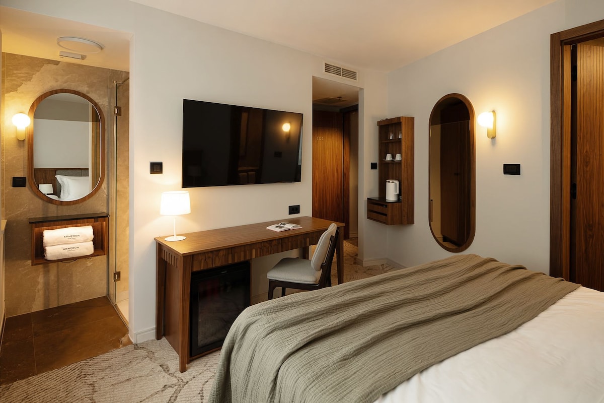 A hotel room with a bed, a desk with a chair, a mirror, a television, and a wardrobe.