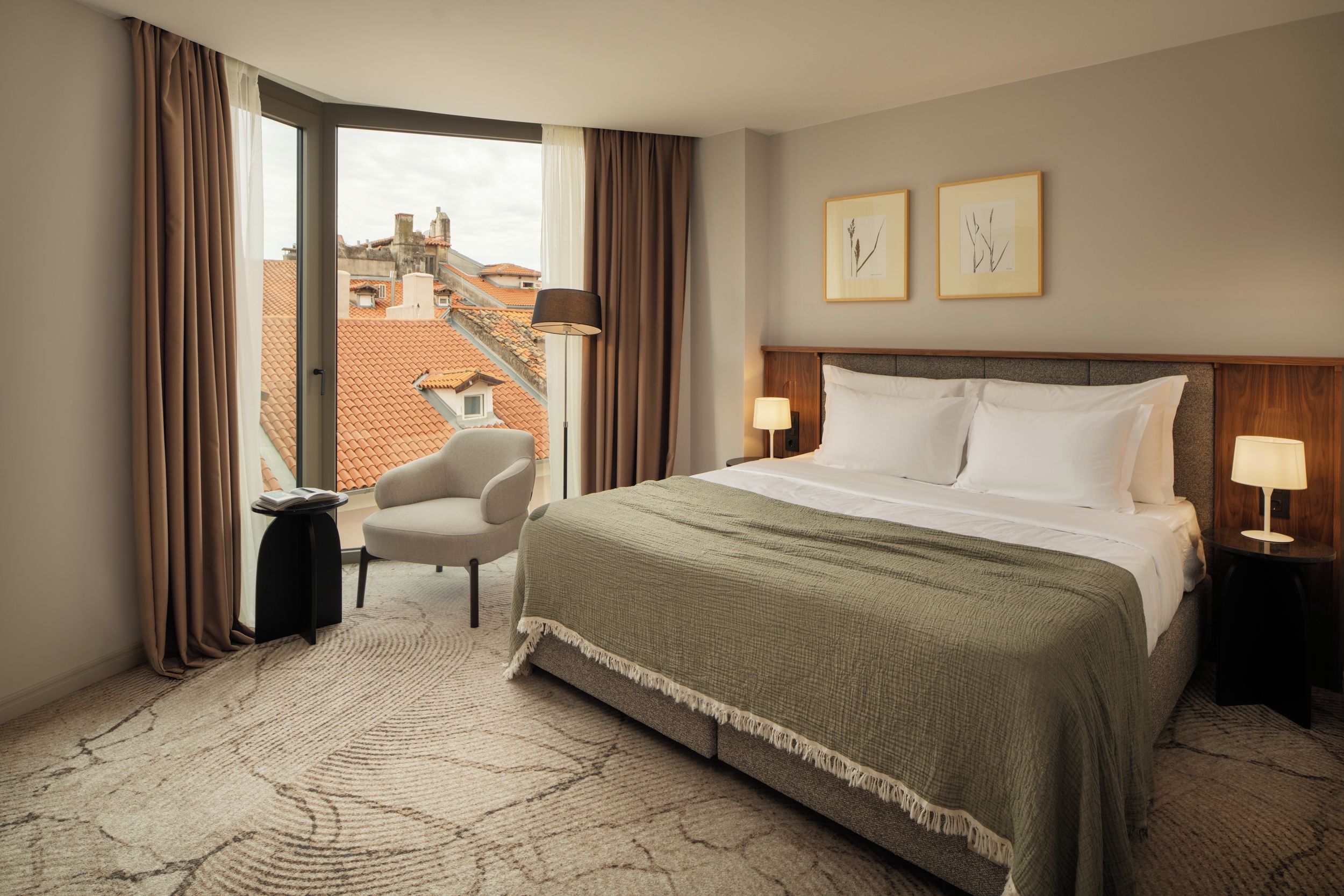 A hotel room with a bed, two bedside lamps, a chair, a window with curtains, and two framed pictures on the wall.