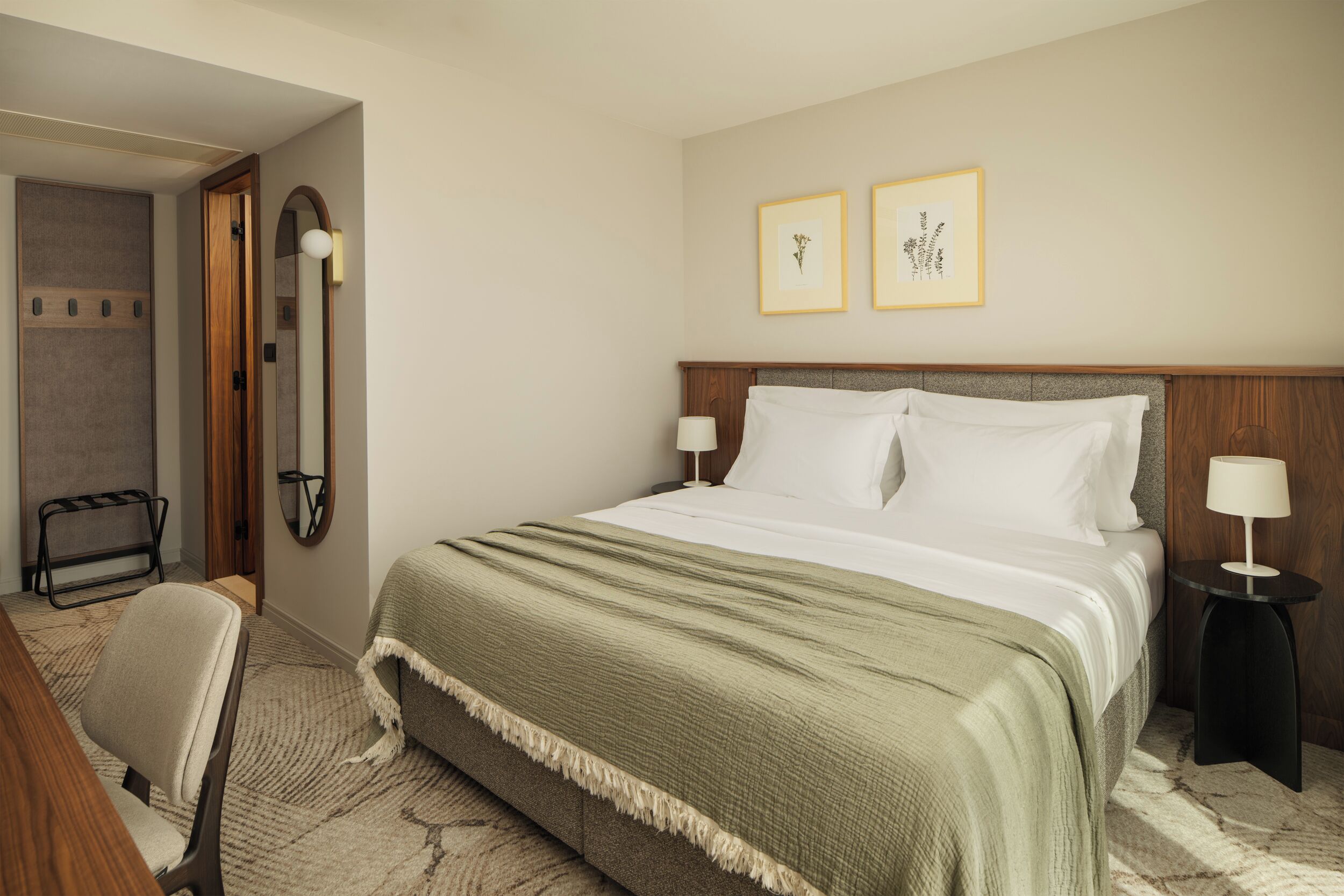 A hotel room with a bed, bedside tables, a chair, a mirror, and artwork on the wall.