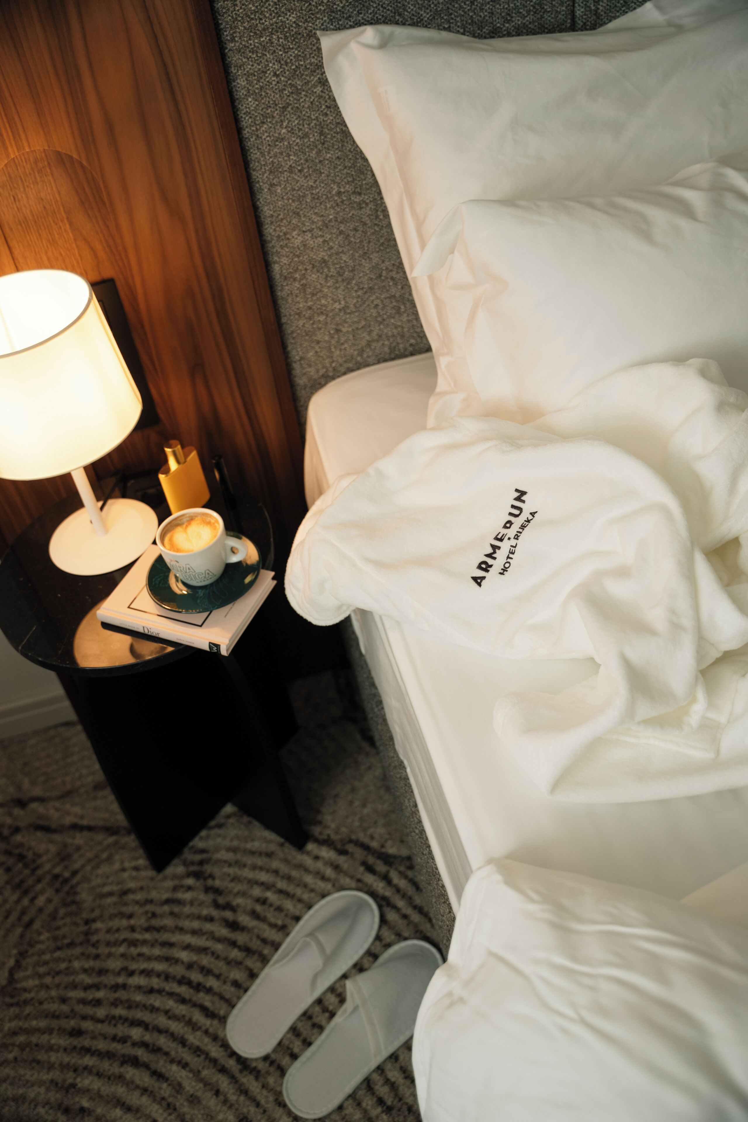 A hotel room with a bed, a nightstand with a lamp, a cup of coffee, and a pair of slippers.
