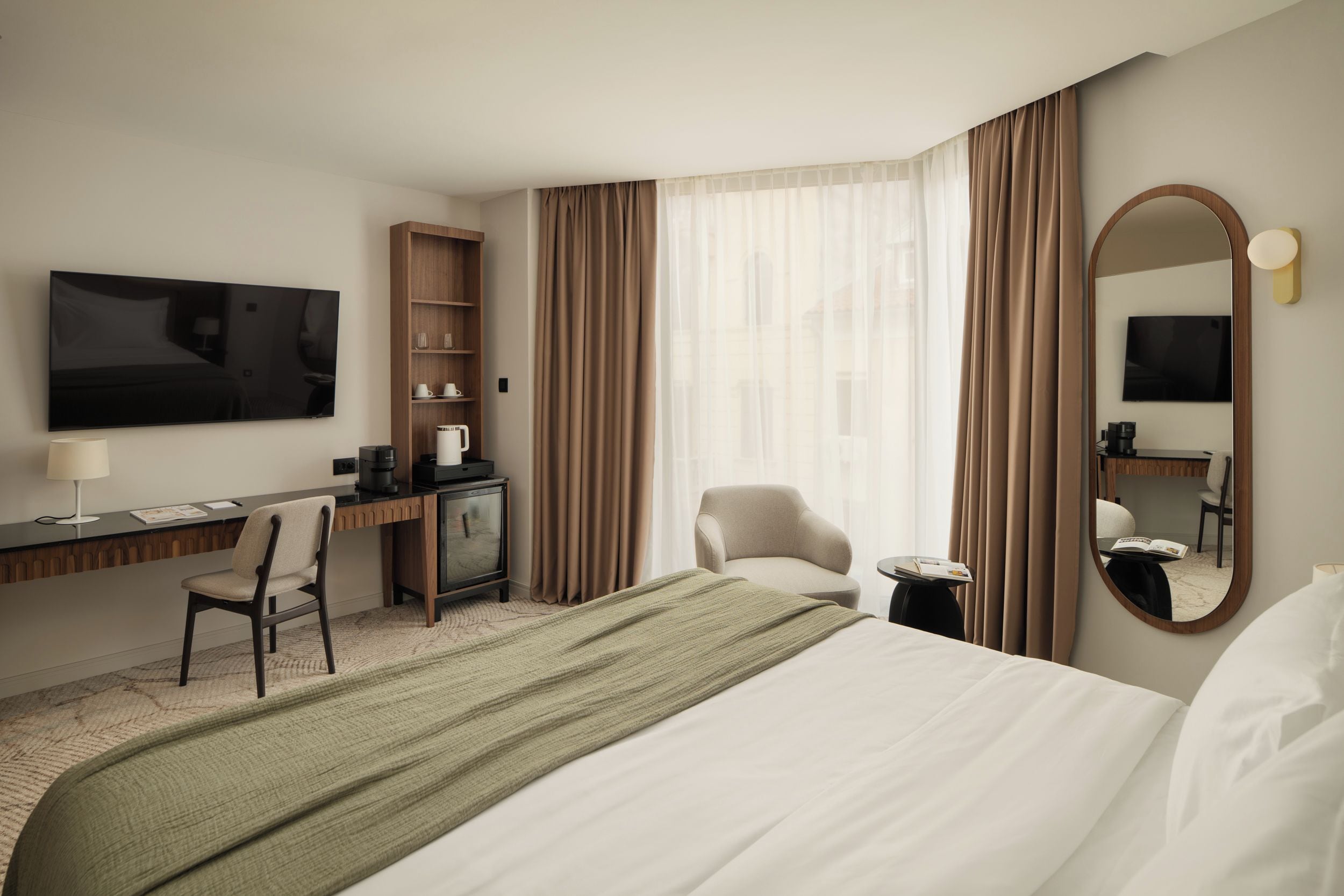 Una habitación de hotel con una cama, un escritorio con silla, televisor, espejo y ventana con cortinas.