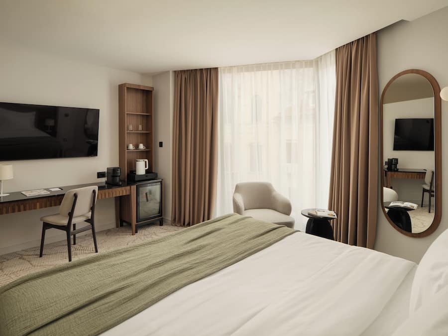 Una habitación de hotel con una cama, un escritorio con silla, televisor, espejo y ventana con cortinas.
