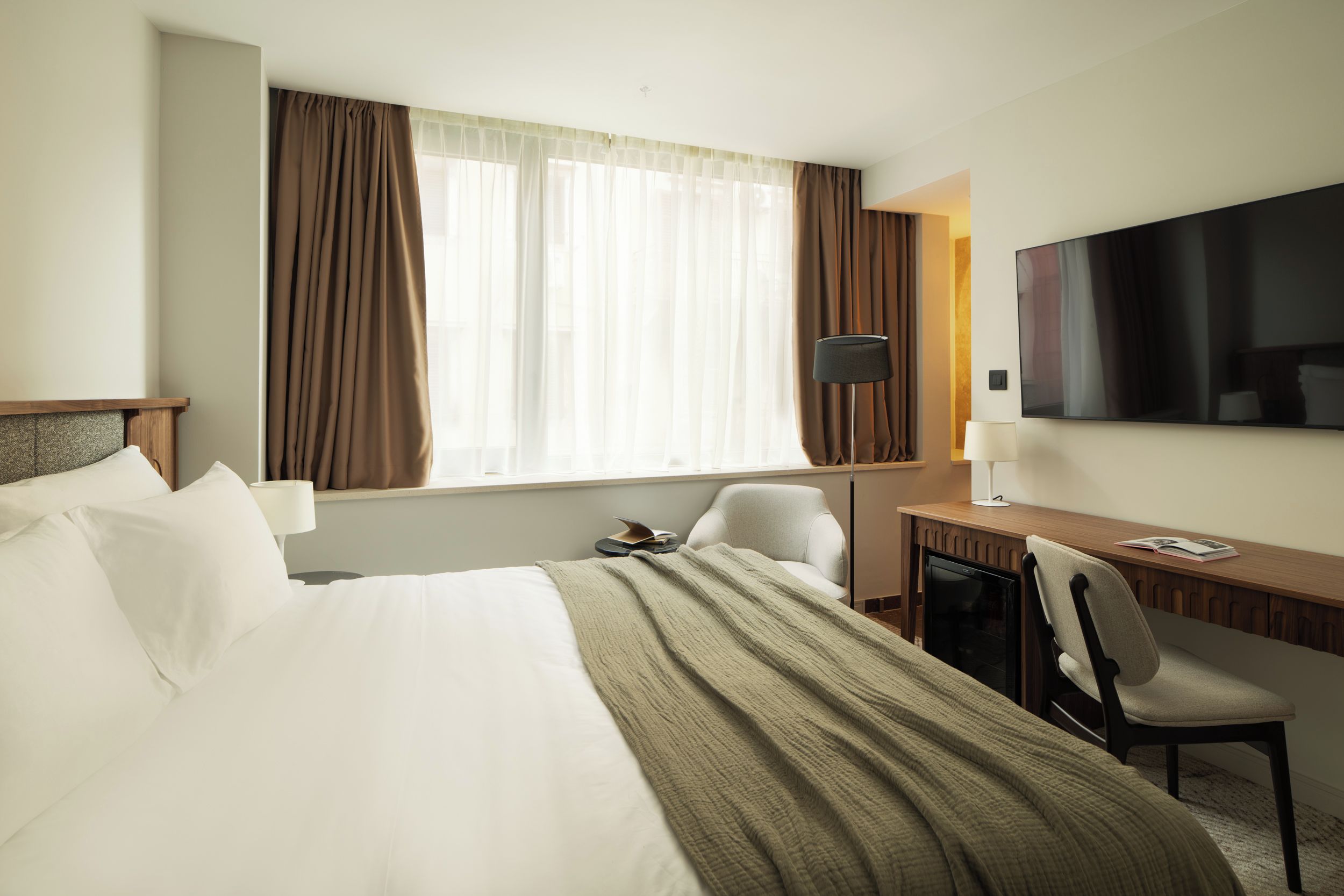 A hotel room with a bed, a desk, a chair, a TV, and a window with curtains.