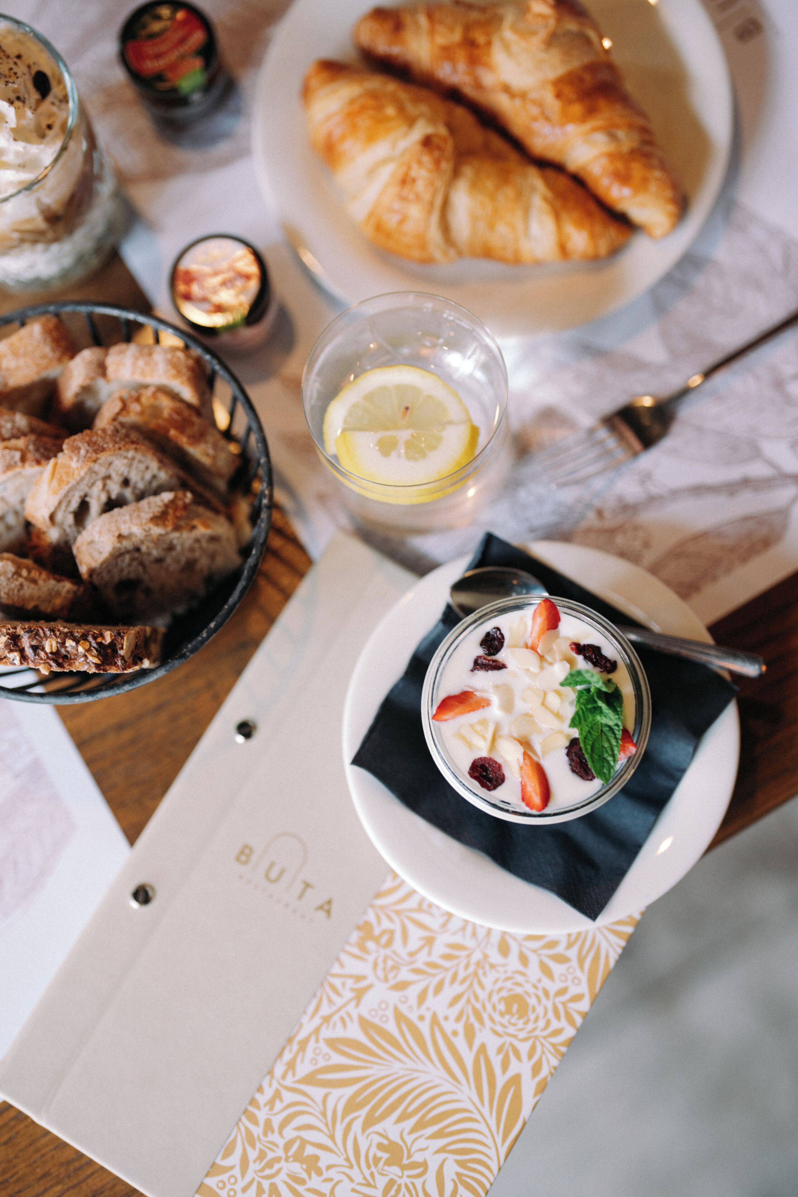 Een eettafel met een bord croissants, een kom yoghurt met fruit en een glas water met een citroenpartje.