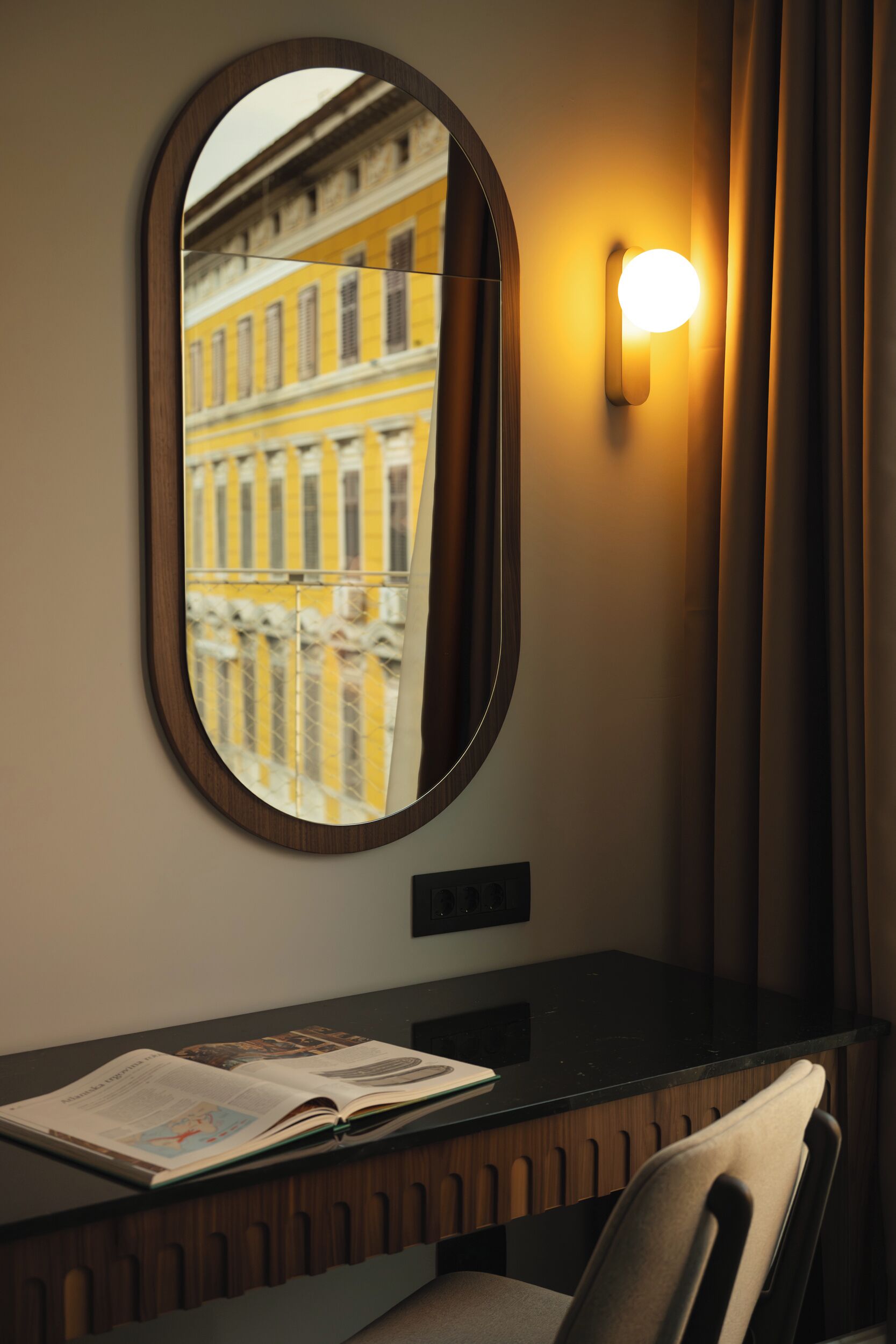 Una stanza con uno specchio che riflette un edificio, una scrivania con un libro e una lampada fissata al muro.