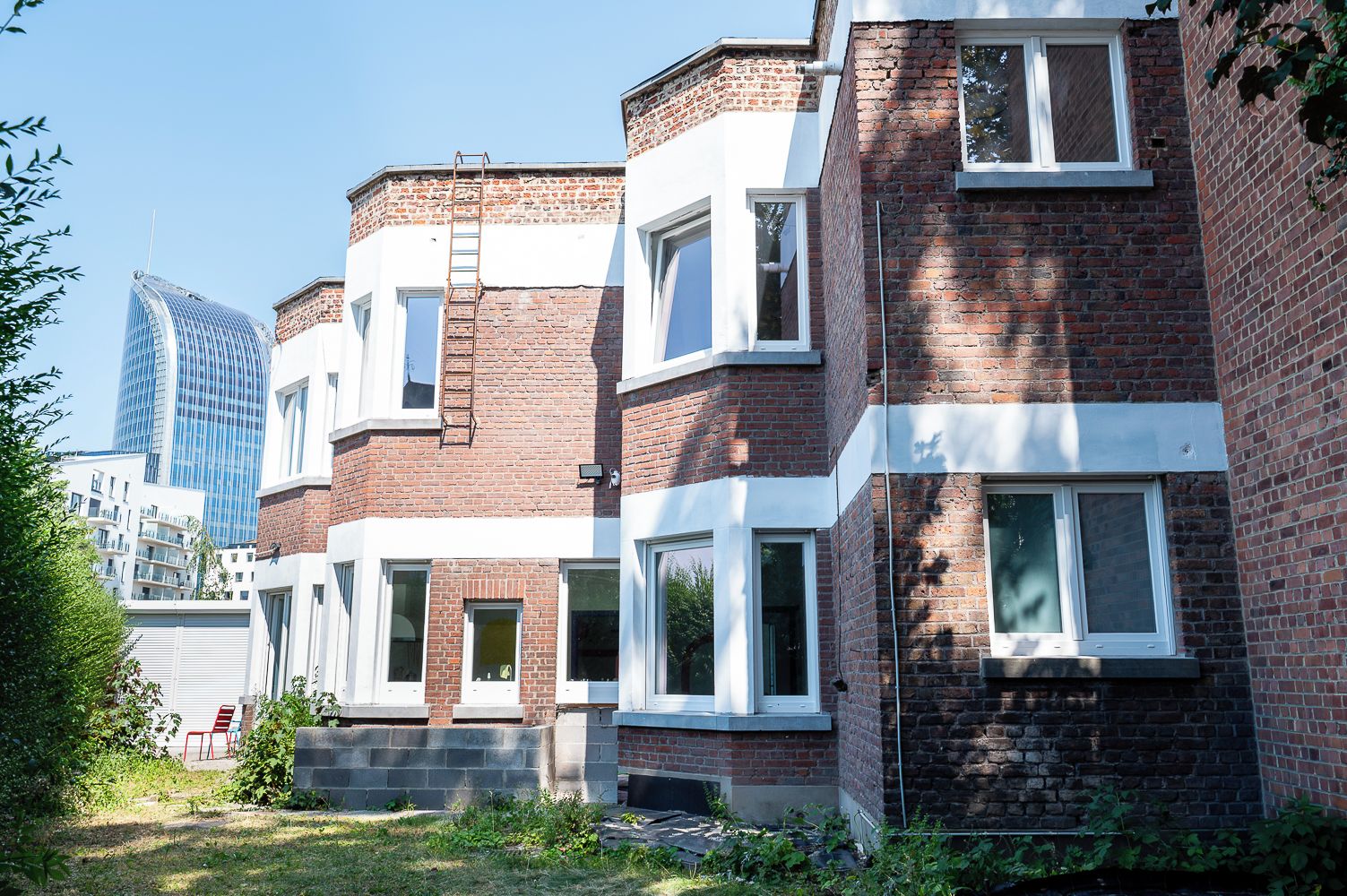 Een rij herenhuisjes van baksteen met witte raamkozijnen, een zichtbare bakstenen schoorsteen en een ladder op het dak.